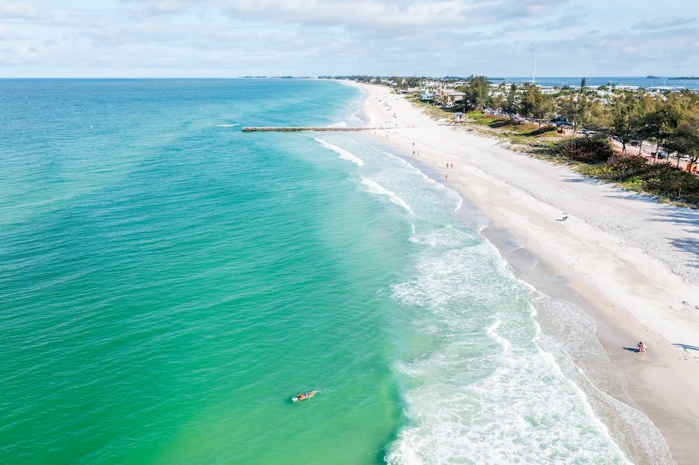Explore the Gulf Island Ferry Stops aerial view of Cortez Beach with turquoise water and a bright sunny sky, a surfer is visible as well as a long concrete pier